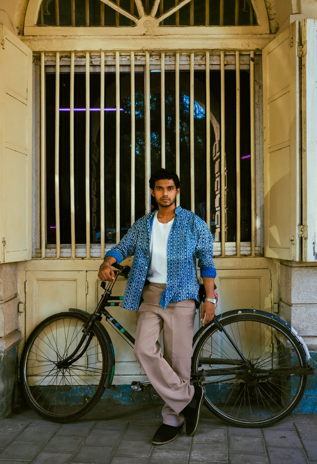 A man wearing a blue patterned shirt, white t-shirt, and beige trousers stands next to a vintage bicycle in front of a window with metal bars.